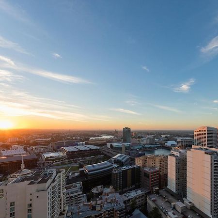 Meriton Suites Kent Street, Sydney Exterior photo The photo shows a city skyline during sunset. In the foreground, there are tall buildings with a modern architectural style, and one building is visibly closer to the camera. The sky is painted with soft hues of orange and blue as the sun sets on the