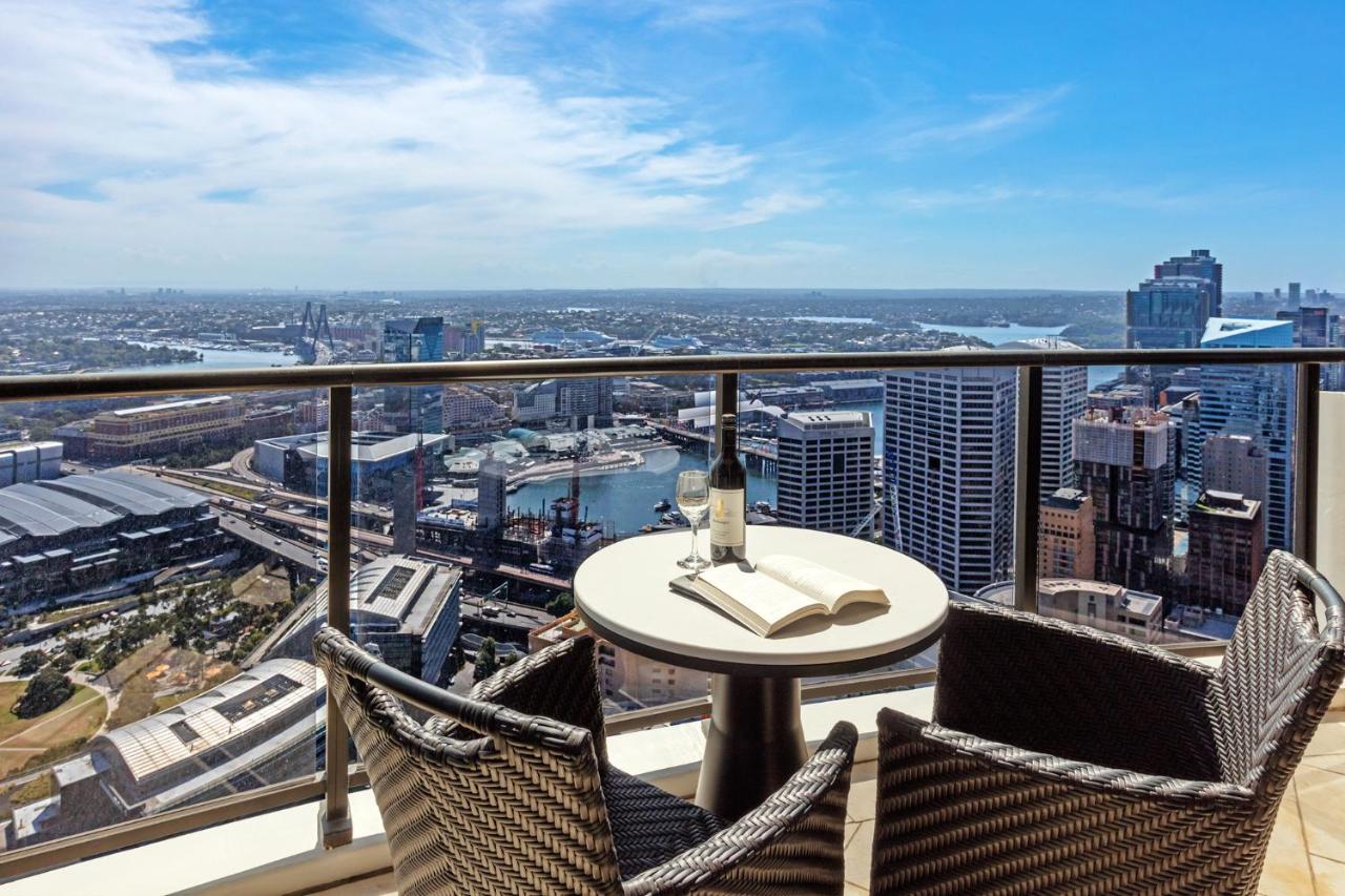 Meriton Suites Kent Street, Sydney Exterior photo The photo shows a beautiful panoramic view from a high balcony. In the foreground, there is a round table with a bottle of wine and an open book on it. Surrounding the table are two comfortable-looking chairs made of woven material. The background fe