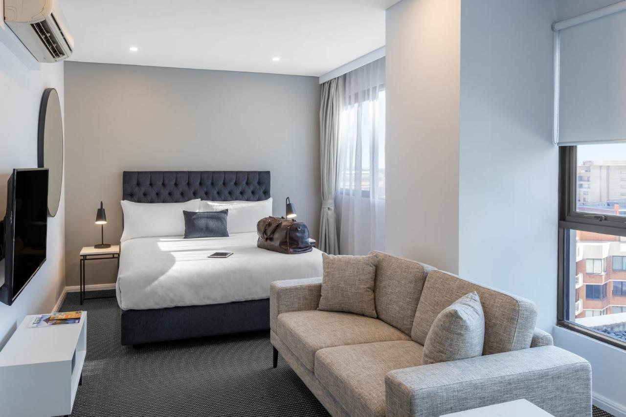 Meriton Suites Kent Street, Sydney Exterior photo The photo shows a modern hotel room. It features a cozy bed with a tufted headboard and white bedding, positioned against a light gray wall. There's a small side table beside the bed, with a lamp on it and a travel bag placed on the bed. A comfortabl
