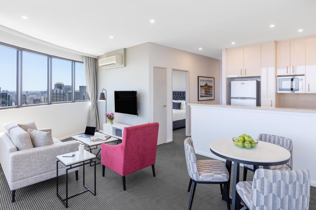 Meriton Suites Kent Street, Sydney Exterior photo The photo shows a modern living space, possibly a hotel room or an apartment. It features a cozy seating area with a light-colored sofa and a pink armchair. There is a small black table beside the sofa and a round dining table with gray chairs, toppe