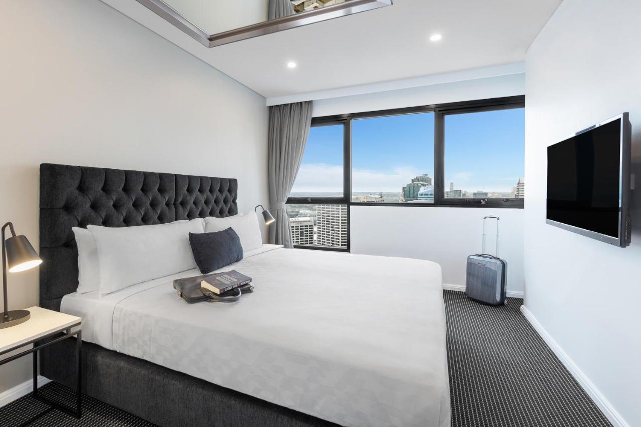 Meriton Suites Kent Street, Sydney Exterior photo The photo shows a modern hotel room featuring a large, neatly made bed with a grey upholstered headboard. There is a decorative cushion on the bed, and a small stack of items, possibly books or magazines, placed at the foot of the bed. Next to the be