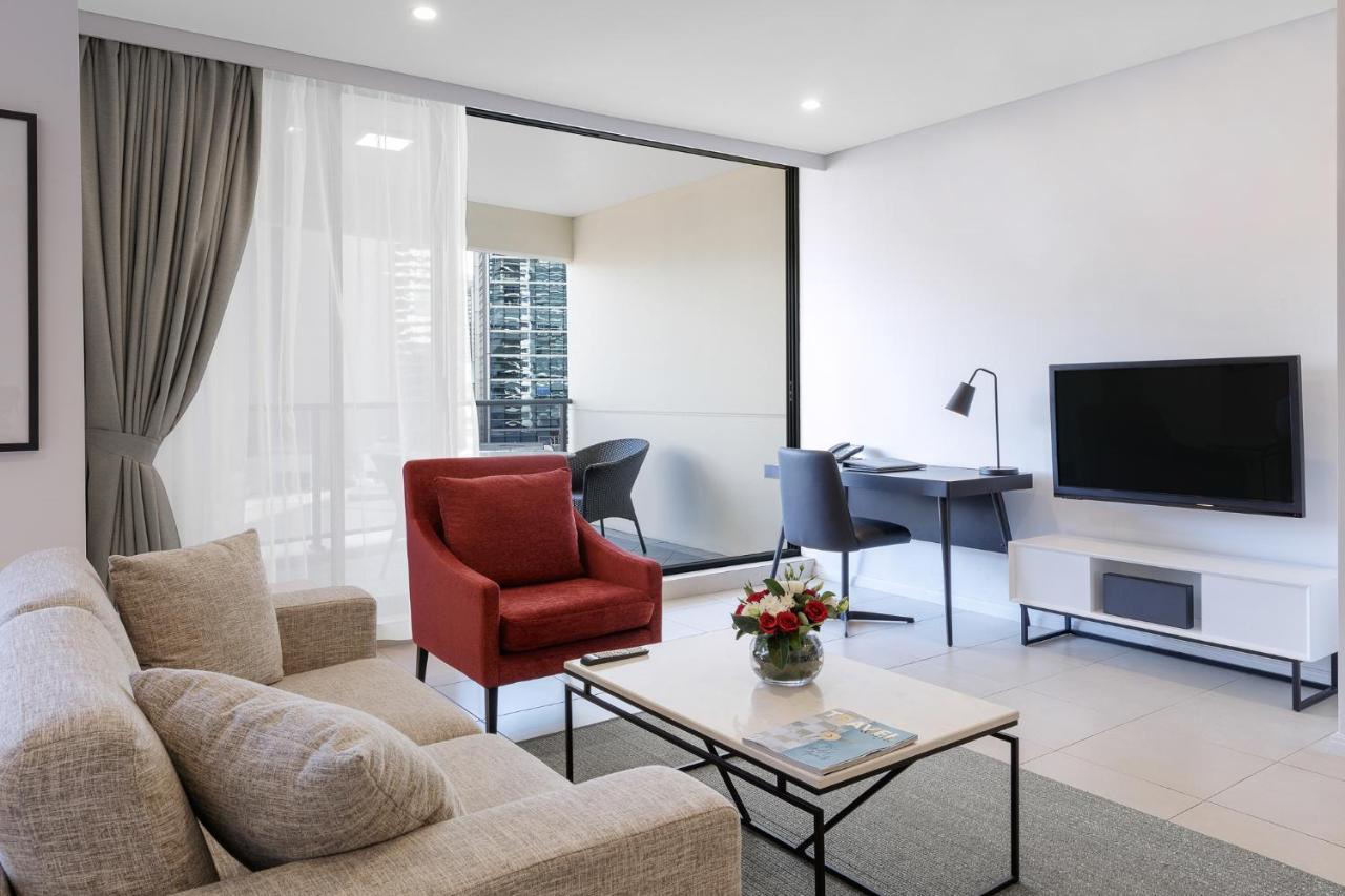 Meriton Suites Kent Street, Sydney Exterior photo The photo shows a modern living space featuring a comfortable cream-colored sofa and a vibrant red armchair. A coffee table sits in front of the sofa, adorned with a small vase of flowers and a magazine. There is a television mounted on the wall oppo